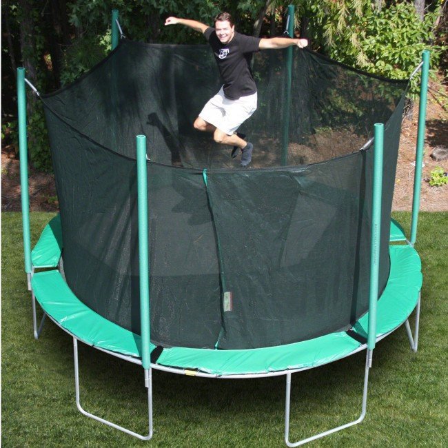 a smiling man in khakis bounces on green, round trampoline with enclosure