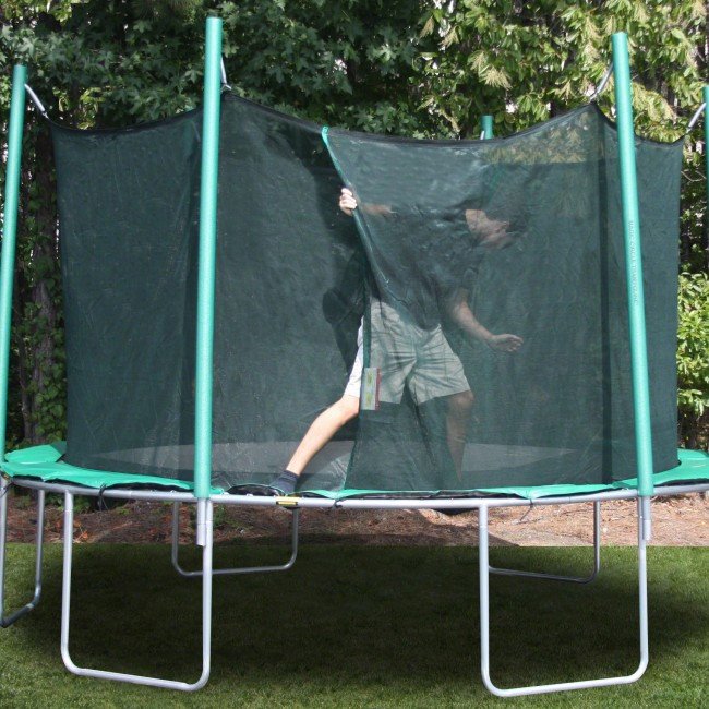 A man in khaki shorts, black shirt, and socks climbs out of a trampoline safety enclosure