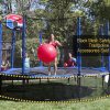 a woman in red bathing suit jumps on a trampoline with black mesh safety skirt while holding a big red ball
