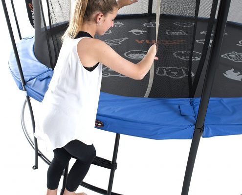 A girl in white tank top and black yoga pants climbs a ladder into a trampoline
